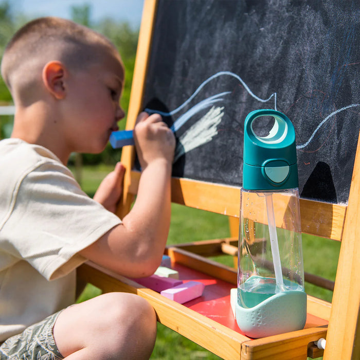 b.box kids tritan drink bottle with teal lid on an art easel beside a child drawing outdoors
