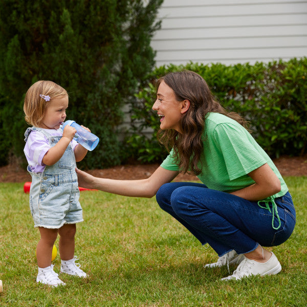 Toddler wearing denim overalls and sneakers drinks from a bottle while standing with a smiling woman outdoors.