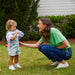 Toddler wearing denim overalls and sneakers drinks from a bottle while standing with a smiling woman outdoors.