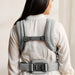 Light grey woven baby carrier harness worn by woman, adjustable straps and padded back