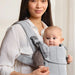 Woman carrying baby in light grey woven baby carrier, front facing, indoors