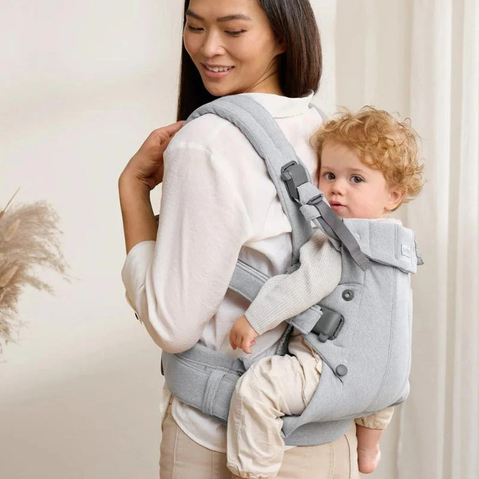 Woman carrying baby in light grey ergonomic baby carrier, cozy indoor setting