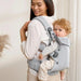 Woman carrying baby in light grey ergonomic baby carrier, cozy indoor setting