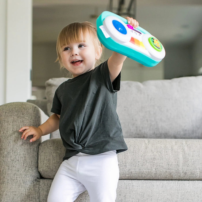Baby Einstein Toddler Jams musical toy held by a smiling toddler standing by a sofa indoors.