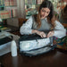 Baby On Board Doudoune diaper bag shown open on a table as a woman organizes baby essentials inside.