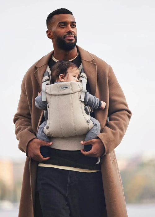 BabyBjorn Harmony baby carrier in beige worn by a man carrying an infant outdoors.