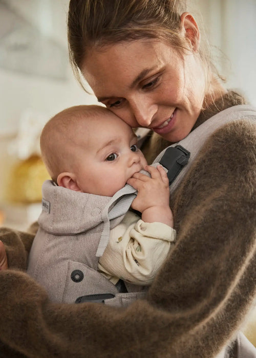 BabyBjörn Harmony baby carrier in beige with a baby held close to an adult's chest indoors.