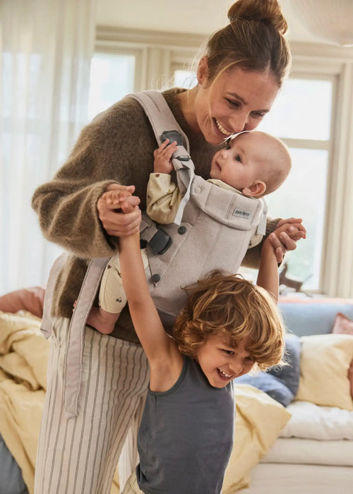 BabyBjorn Harmony baby carrier in beige with a baby being carried indoors by a smiling adult.