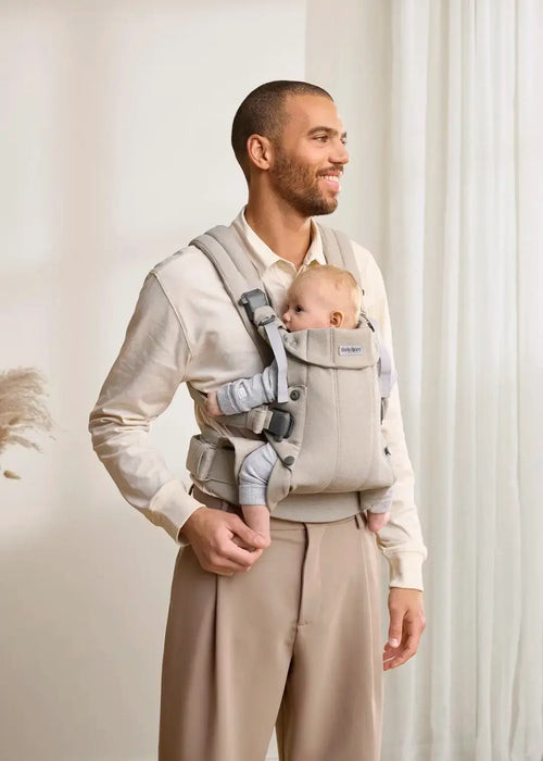 BabyBjörn Harmony baby carrier beige with baby facing inward, worn by an adult in a home setting.
