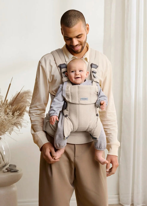 BabyBjorn Harmony baby carrier in beige worn by a smiling adult holding a happy baby indoors.