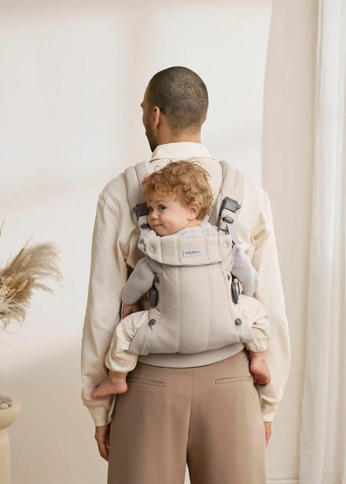 BabyBjorn Harmony baby carrier in beige holding a baby on an adult's back indoors.
