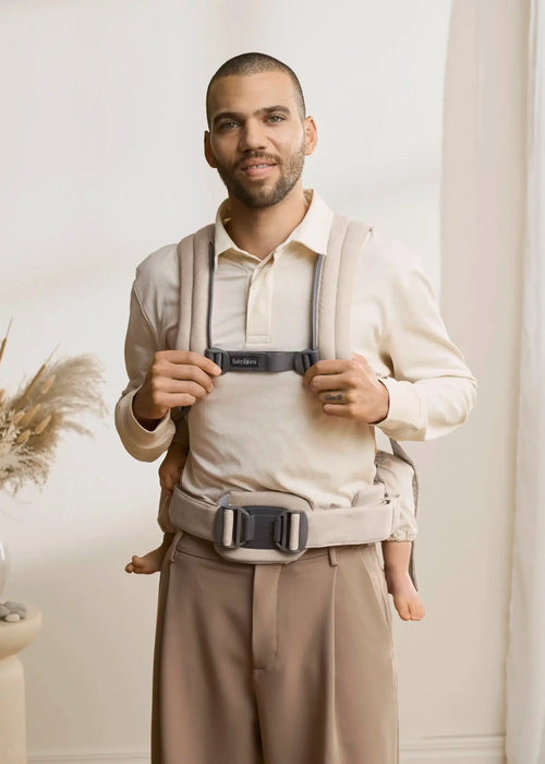 BabyBjorn Harmony baby carrier in beige worn by an adult holding a baby indoors.
