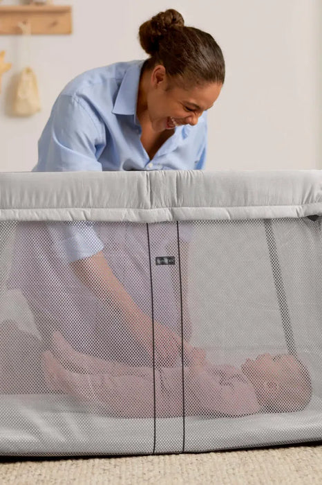 BabyBjorn Play Yard Light Silver with a smiling woman tending to a baby inside the mesh-sided play yard.