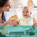 Biodegradable disposable baby bibs shown on a smiling baby in a high chair being fed in a kitchen.
