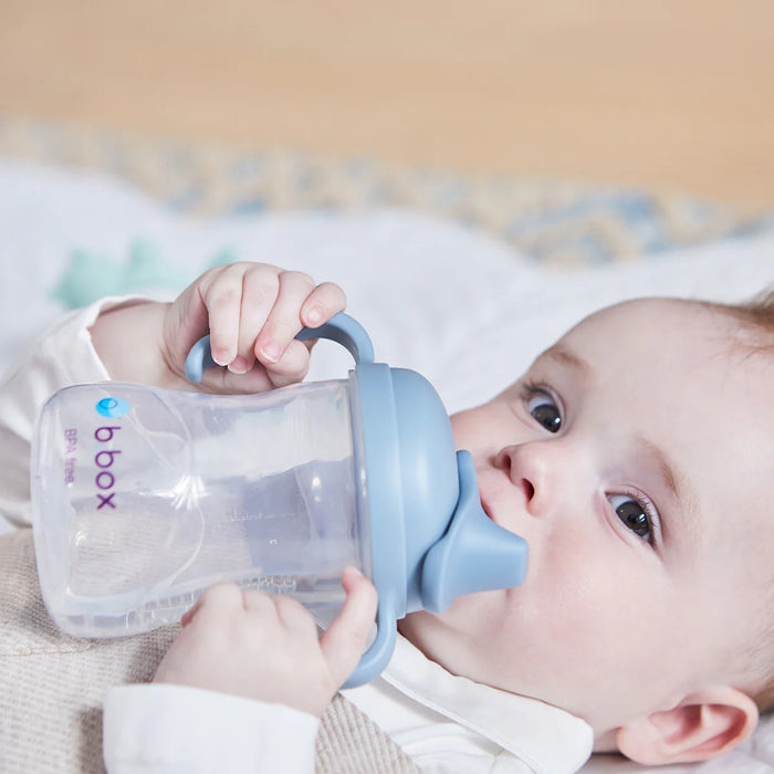 bbox silicone sippy cup held by baby lying on a blanket, sipping from blue lid.