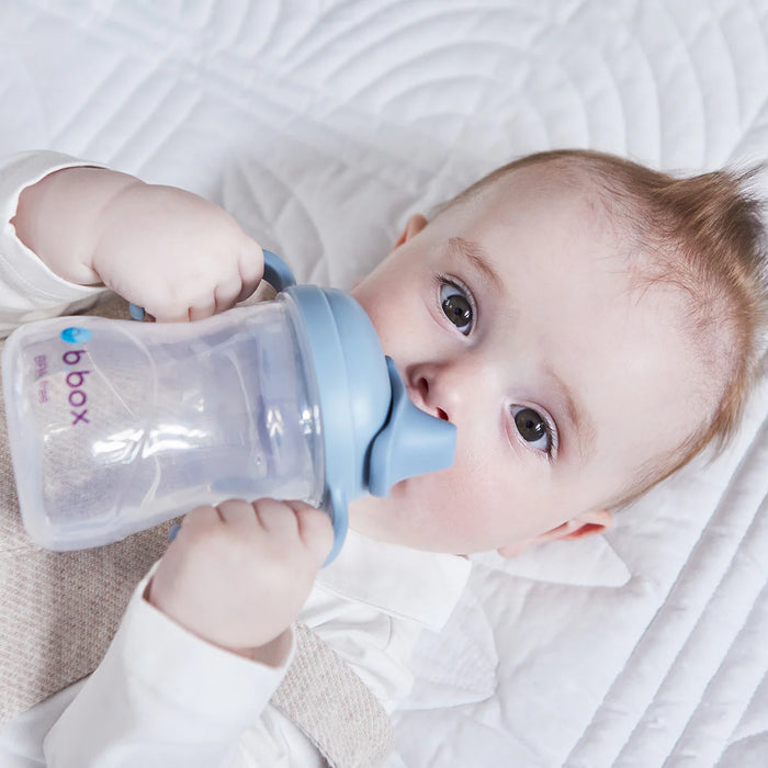 b.box silicone sippy cup in blue held by baby lying on a white quilted blanket.