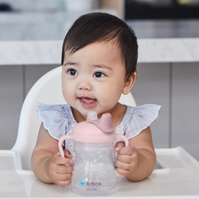 bbox silicone sippy cup in blush held by a baby sitting in a high chair at a table.