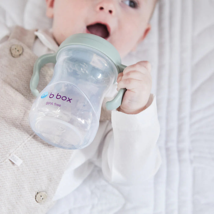 b.box silicone sippy cup in sage color held by baby lying on a soft white blanket.