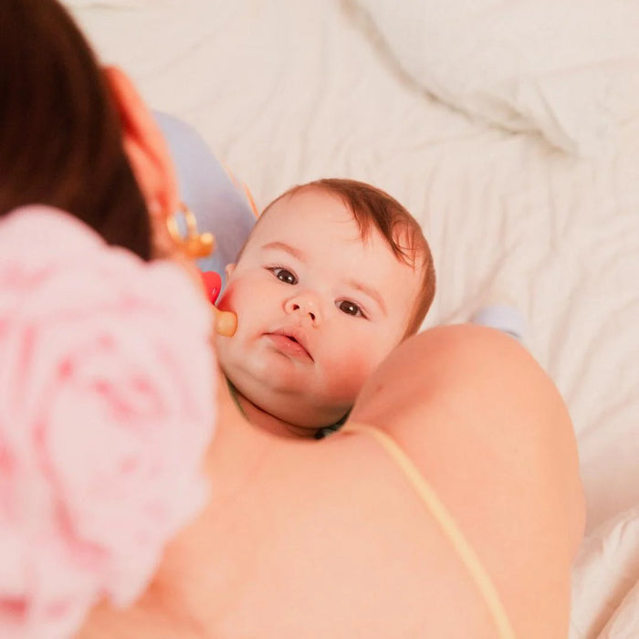 A baby lying on a bed looking up, cradled in an adult's arms, with a soft focus background.