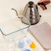 Bibs glass baby bottle with latex nipple and accessories on a countertop near kettle and towel.