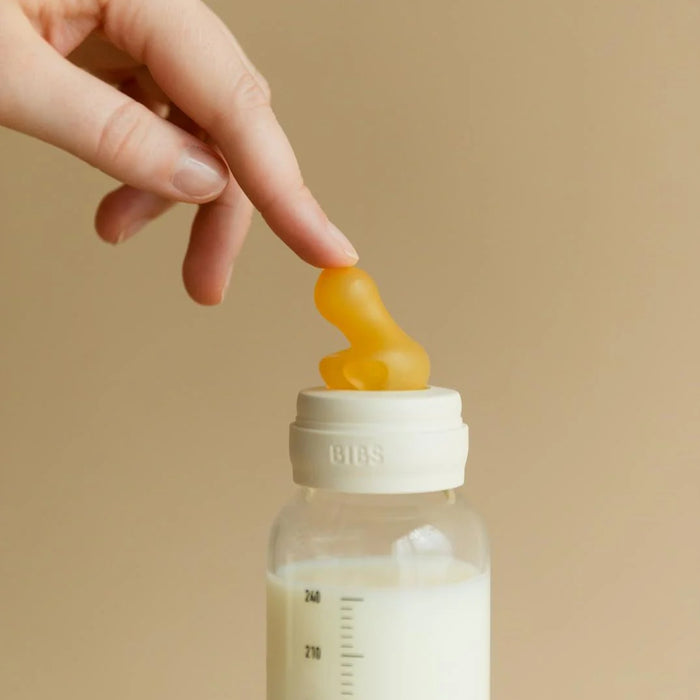 BIBS glass baby bottle with ivory cap and latex nipple, shown with a hand touching the nipple.