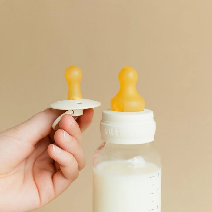 BIBS glass baby bottle with latex nipple and pacifier held by hand against beige background.