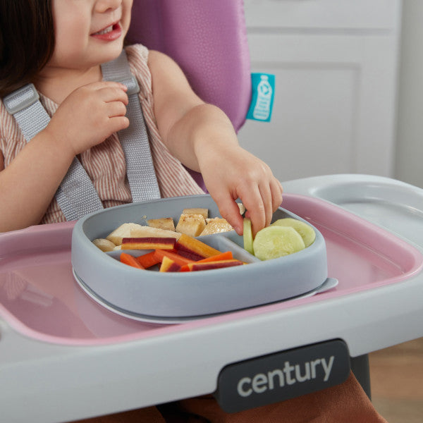 A toddler in a high chair reaching for food on a divided silicone plate during mealtime.