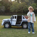 Ride-on toy car with open door shown outdoors on grass beside a smiling child in casual clothes.