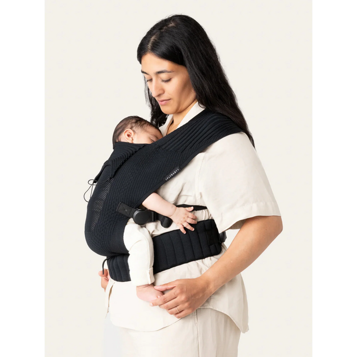 Flybaby 3D baby carrier in black shown worn by an adult holding a sleeping baby close to their chest.