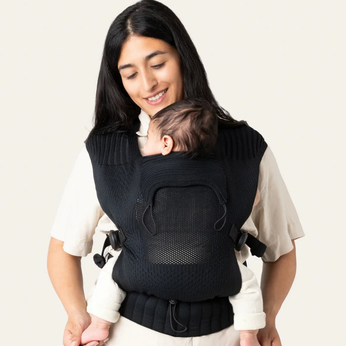 Flybaby 3D Baby Carrier in black shown worn by a parent carrying a baby on their chest indoors.