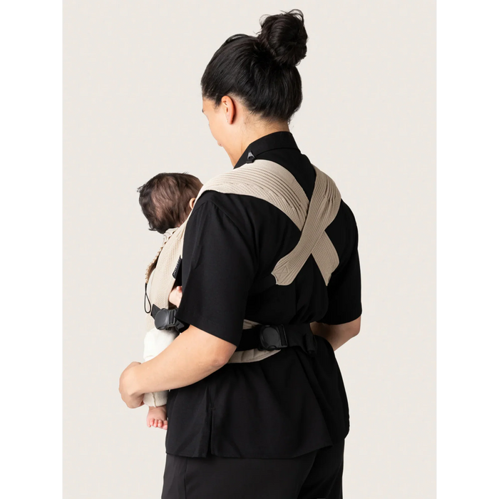 Flybaby 3D baby carrier with beige straps shown worn by a woman holding a baby from the back view.