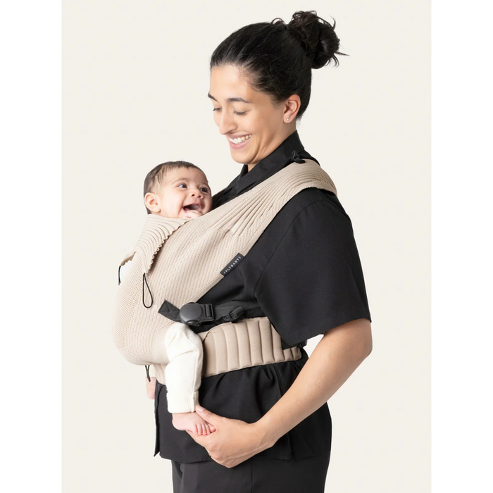 Flybaby 3D baby carrier in beige with a smiling baby carried by an adult wearing black.