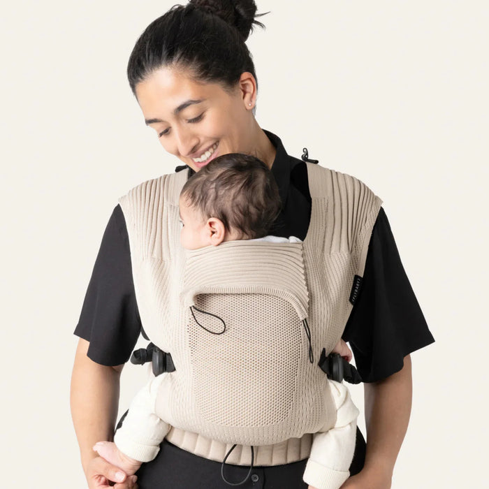 Flybaby 3D baby carrier in beige shown worn by an adult holding an infant on their chest.