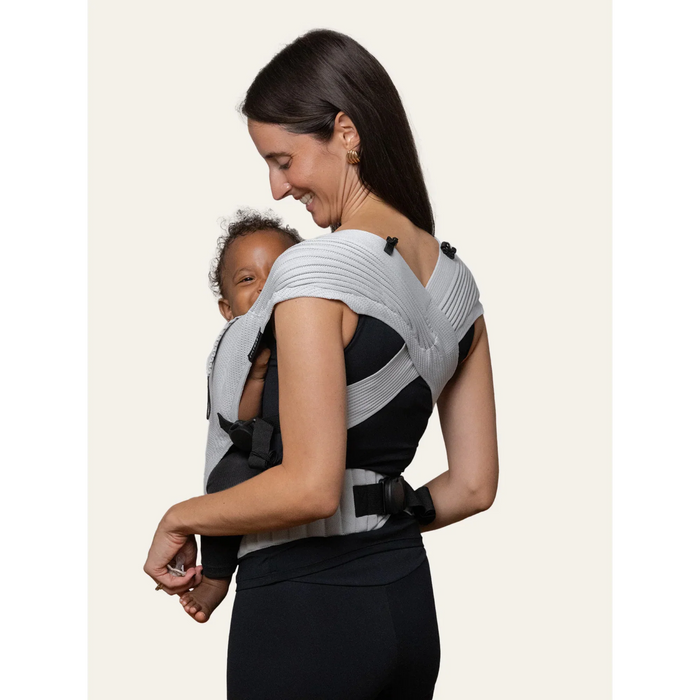 Flybaby 3D Baby Carrier in grey worn by a woman carrying a smiling baby on her front.