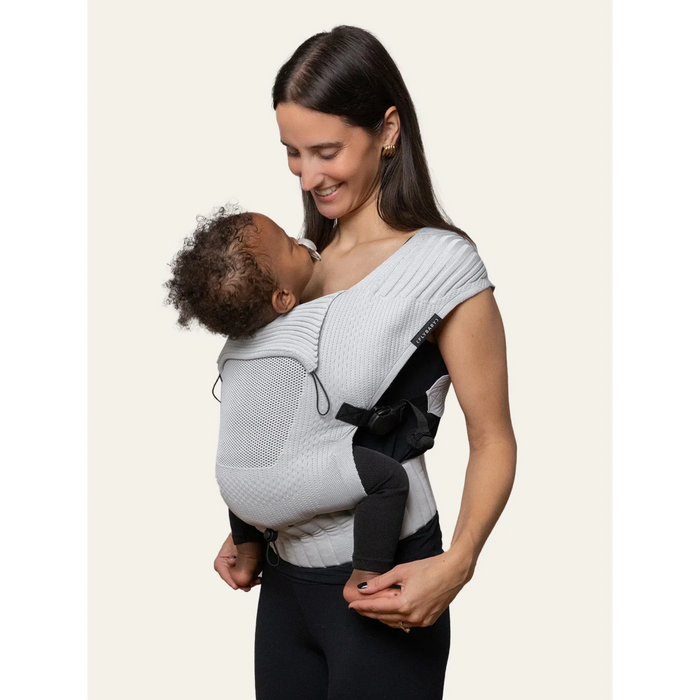 Flybaby 3D baby carrier in light grey, worn by a smiling woman holding an infant close to her chest.