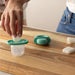 Frida Baby Push Pop Feeder being filled with ice by a hand on a wooden countertop.