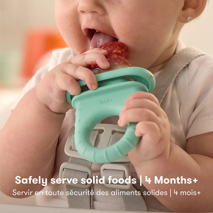 Frida Baby Push Pop Feeder held by a baby in a high chair, baby chewing on the feeder filled with fruit.