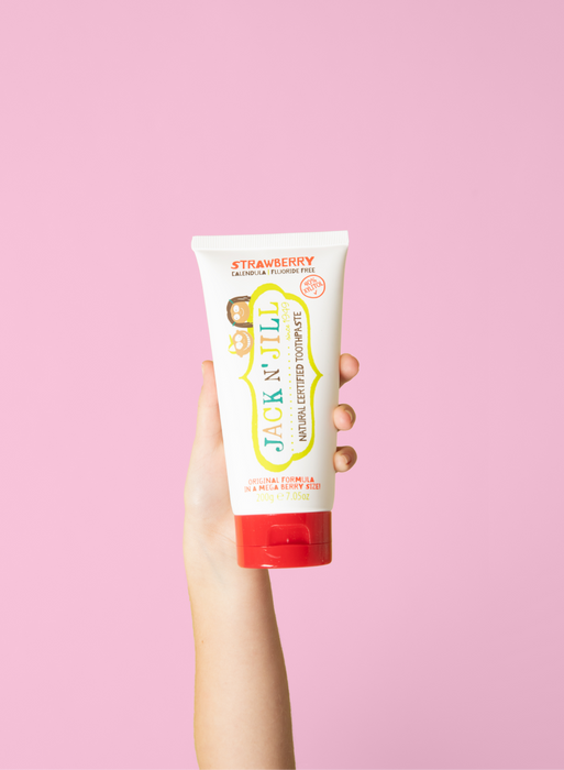Jack N' Jill Strawberry Natural Toothpaste held in a hand against a pink background.