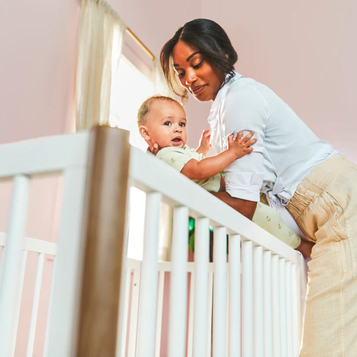 Little Seeds Laurel Island Convertible Crib with mother placing baby inside in a softly lit nursery.