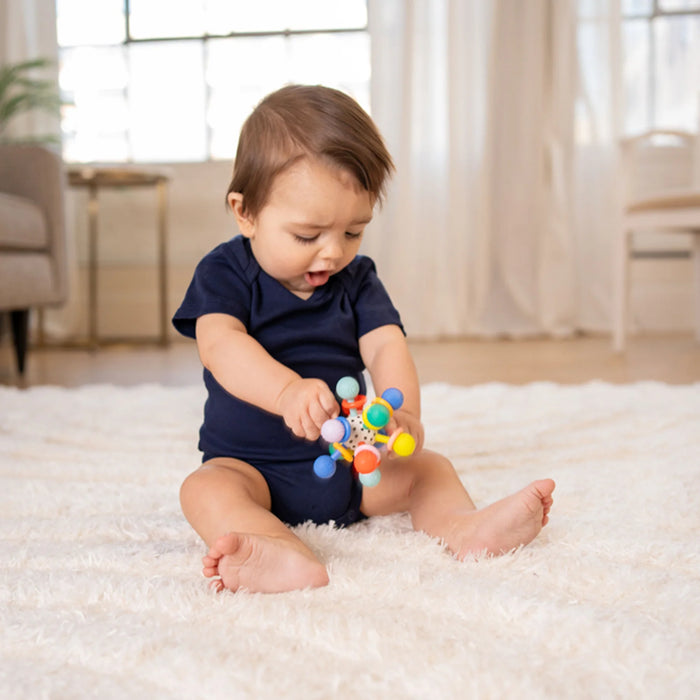 Manhattan Toy Atom Colorpop Rattle Teether held by a baby sitting on a white rug indoors.