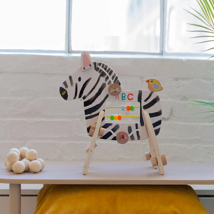 Manhattan Toy Safari Zebra wooden activity toy with colorful beads on a table near a window.