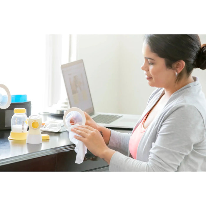 Medela Quick Clean Breast Pump Wipes used by a woman cleaning pump parts at a desk with a laptop.