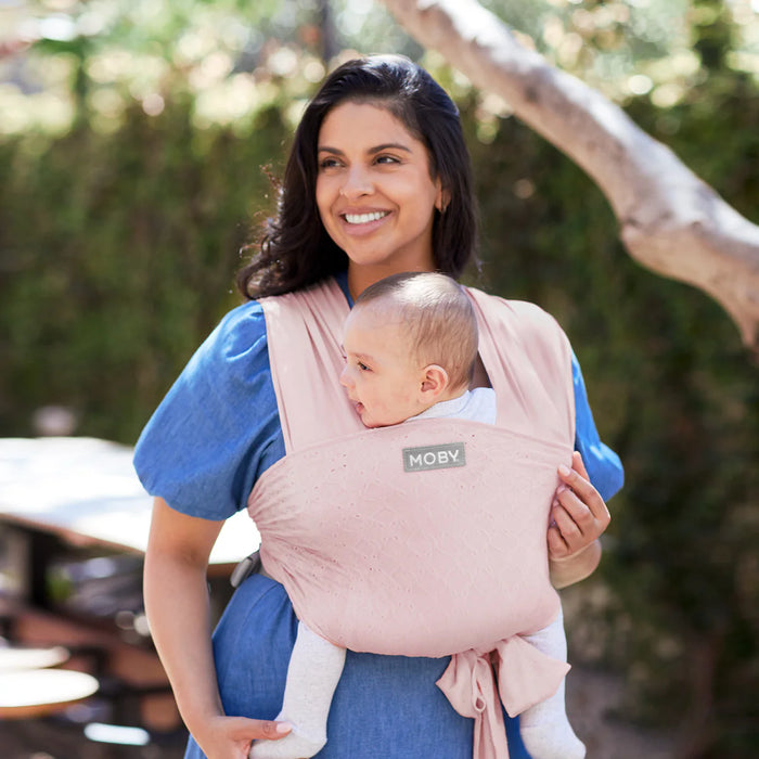 Moby Easy Wrap Baby Carrier in rose, shown worn by a woman carrying a baby outdoors.
