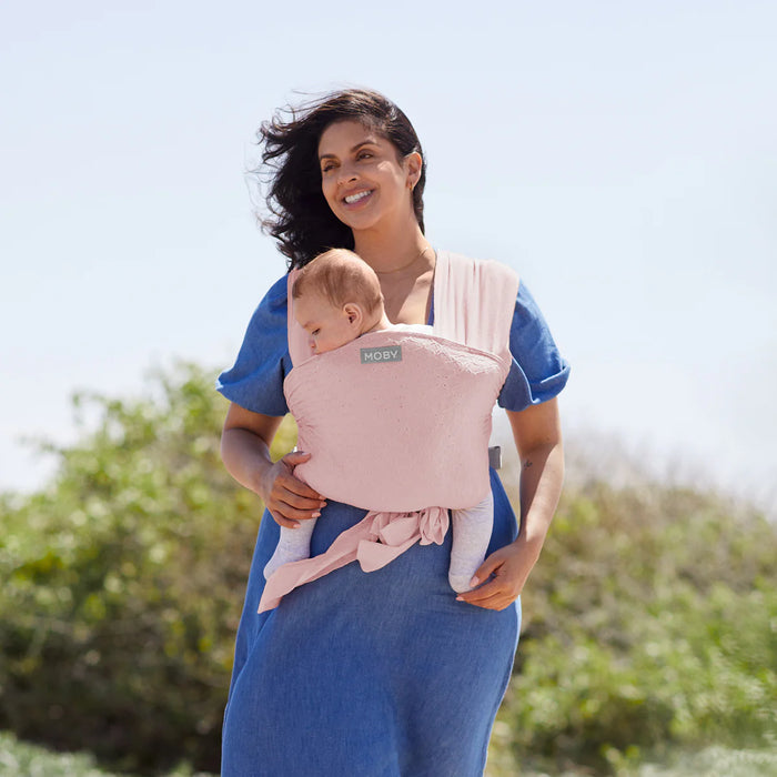 Moby Easy Wrap baby carrier in rose pink shown worn by a woman carrying a baby outdoors.