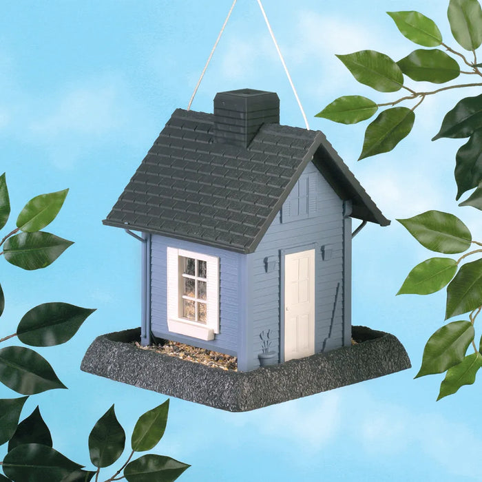 Blue cottage-shaped bird feeder with white door, hanging outdoors, leafy branches nearby