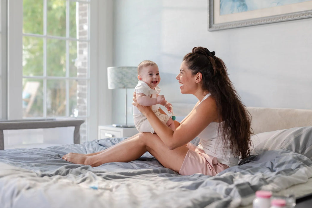 Mother sitting on a bed holding a smiling baby in a sunlit modern bedroom