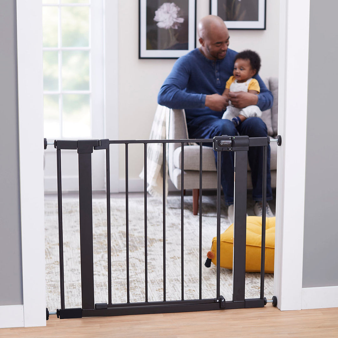 Black walk-through baby safety gate installed in doorway, living room with adult and child in background