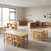 A bright, modern, and empty daycare classroom featuring high-quality solid wood children's furniture. In the foreground are a rectangular activity table with six matching chairs and a square table with four chairs. The background shows low wooden storage shelves filled with toys and educational decor, illuminated by natural sunlight from large windows.