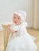 Warm Wendy off-white lace baptism dress shown on a baby standing indoors near a wooden stool.