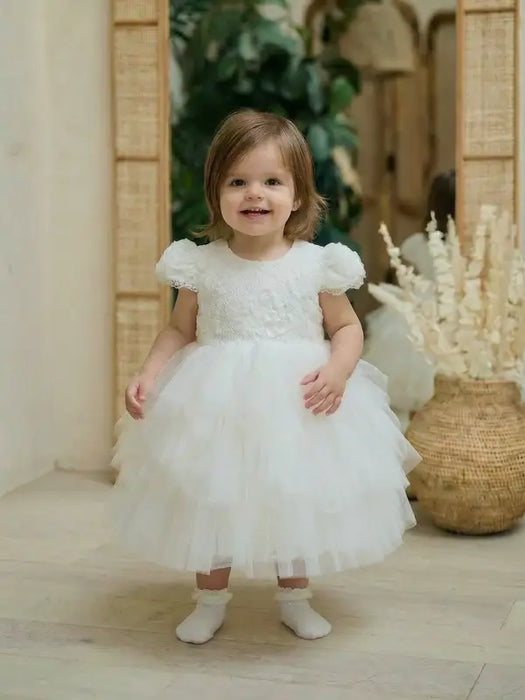 Warm Wendy Off-White Lace Baptism Dress worn by a toddler standing indoors on a light wood floor.